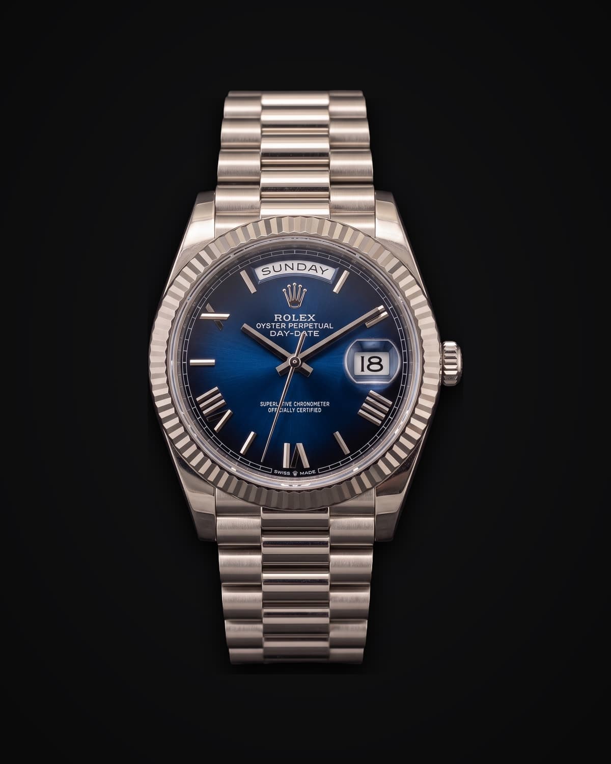 White gold watch with a blue dial fading to black at the edge, Roman numerals, a day window, a fluted bezel and a President bracelet