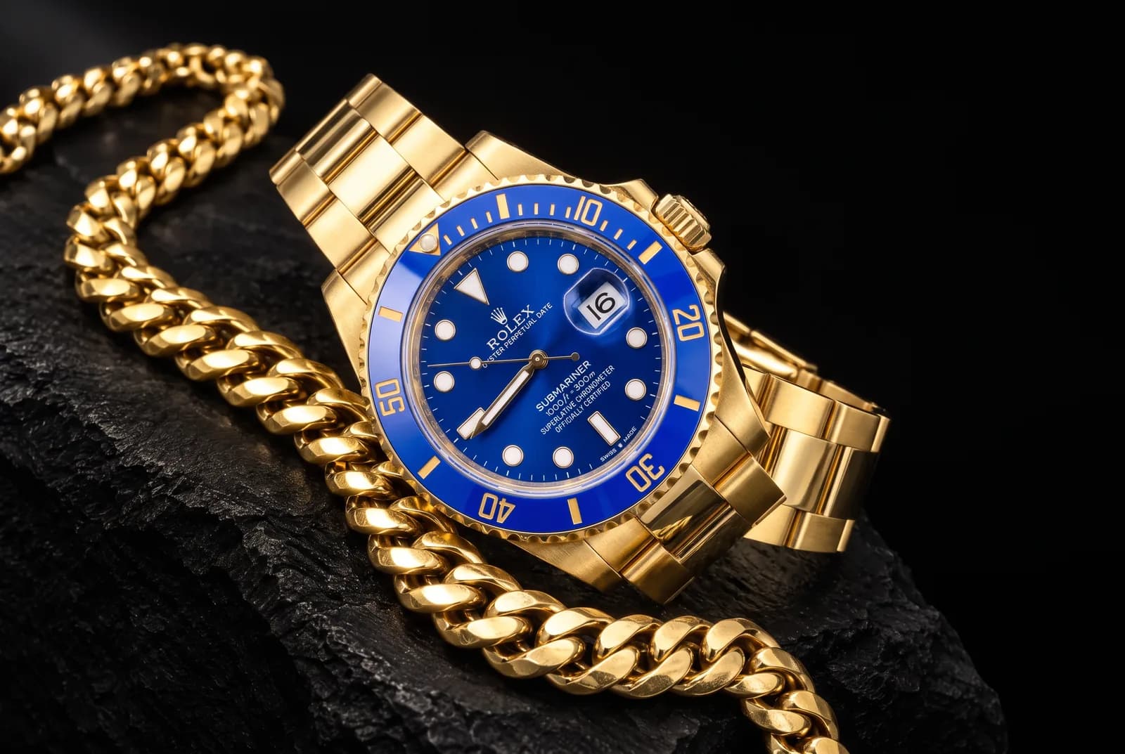 A yellow-gold Rolex Submariner with a blue dial and bezel lying across a heavy gold Cuban link chain, on black stone