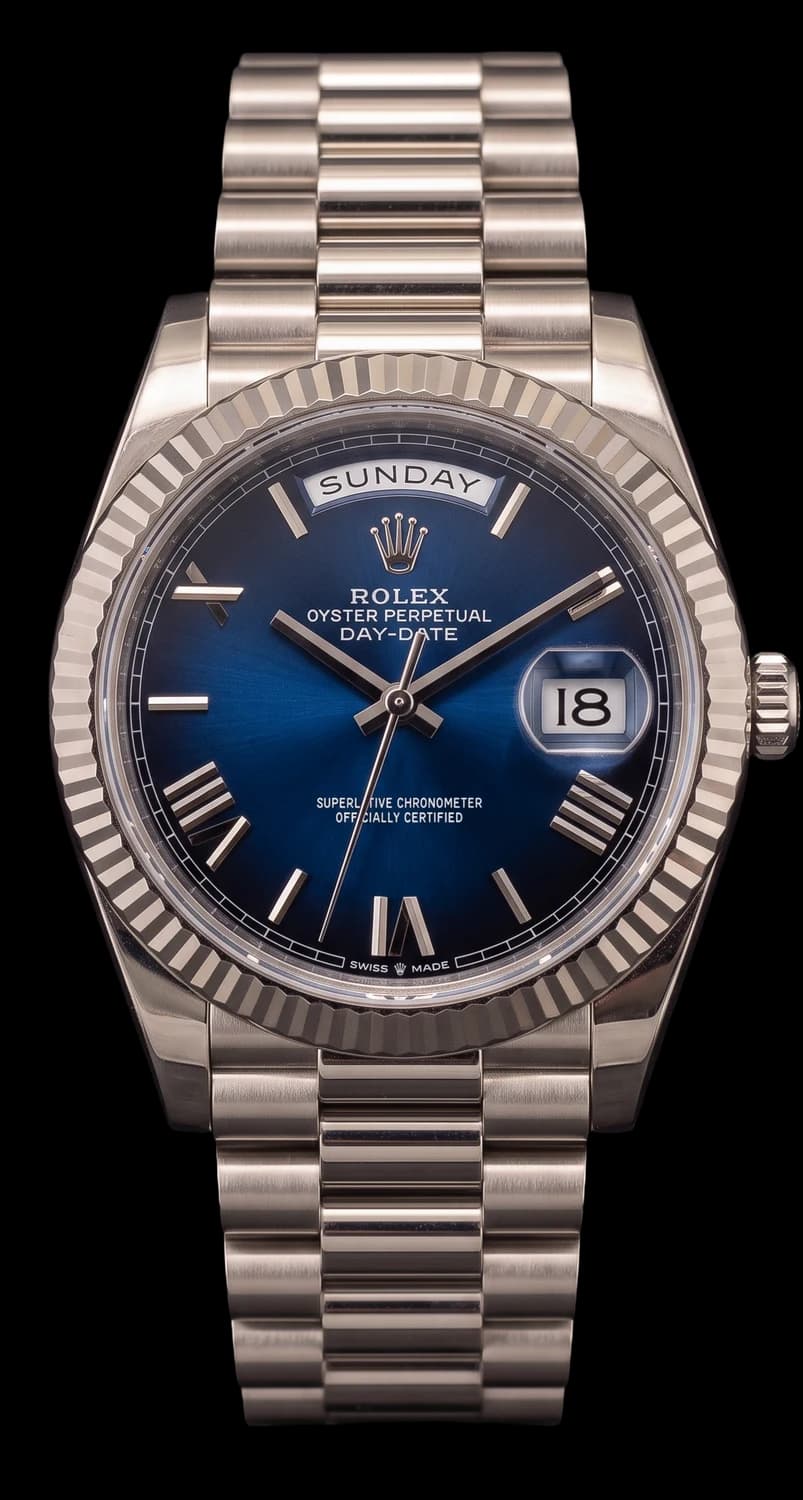 White gold watch with a blue dial fading to black at the edge, Roman numerals, a day window, a fluted bezel and a President bracelet